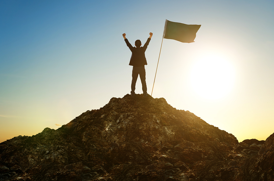 Man standing at the top of a hill with his hands raised in victory with a flag beside him